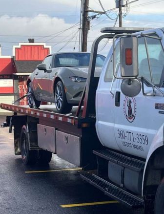 A white flatbed tow truck from Lucky Lab Towing LLC is parked in a lot, transporting a silver sports car near a red commercial building—demonstrating prompt and professional towing services in busy urban areas.
