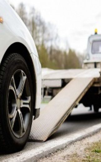 A close-up view of a white car positioned in front of a lowered flatbed ramp, ready to be loaded onto a tow truck parked on the roadside—illustrating the start of a professional vehicle towing operation.