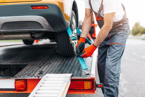 A towing professional securing a yellow car with a blue strap onto a flatbed truck, showcasing emergency towing services on the roadside.
