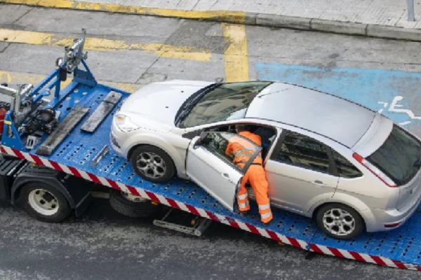 A tow truck operator in an orange uniform securing a silver car onto a flatbed tow truck during daylight, providing emergency towing services on a city street near a handicap parking space.