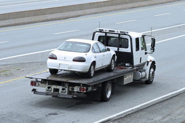 A white sedan is transported on the back of a heavy-duty flatbed tow truck along a multi-lane highway, representing efficient and secure Flatbed Towing services for roadside assistance and vehicle recovery.