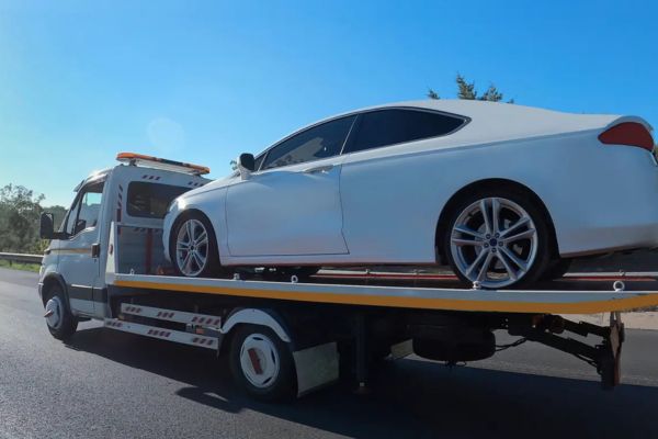 A white luxury car is securely loaded on a flatbed tow truck driving on a sunny highway, demonstrating reliable and damage-free Flatbed Towing services for high-value or immobile vehicles.