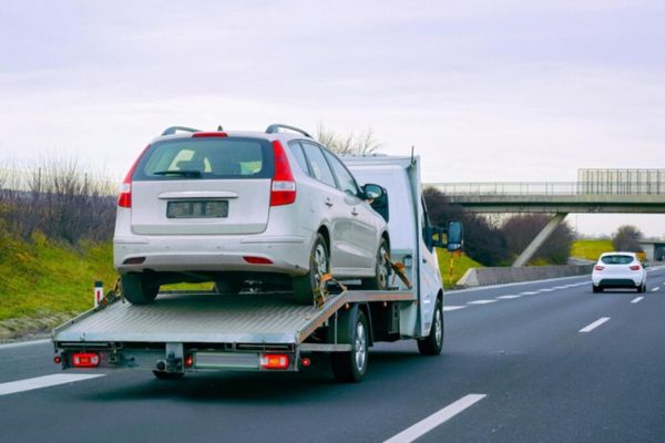 A flatbed tow truck carries a white car along a highway under an overpass, showcasing dependable Local and Long-Distance Towing services for vehicles in transit across short and long routes.