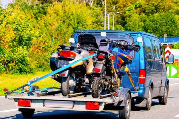 A blue van tows a trailer loaded with multiple motorcycles securely fastened with straps, illustrating reliable and efficient Motorcycle Towing services for group transport or long-distance hauling.